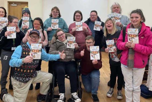 A cheerful group of people pose indoors, each holding a colourful leaflet or programme, smiling towards the camera. The setting appears to be a community or workshop space with wooden floors and a plain wall.