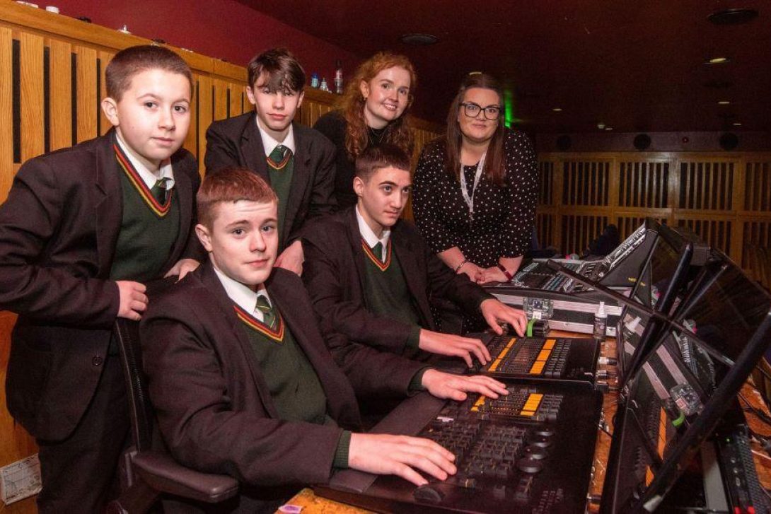 Group of schoolboys in uniform seated and standing at a professional lighting and sound control desk inside a theatre booth. Two female staff members stand behind them, smiling, as the students engage with the technical equipment.