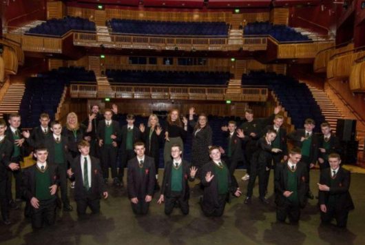 A group of school students in uniform pose on stage inside the Millennium Forum auditorium, with raised arms and the theatre's seating tiers visible behind them.