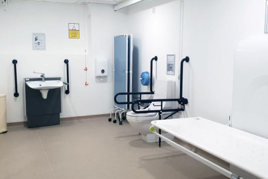Accessible Changing Places facility with height-adjustable bench, support rails, accessible toilet and sink, and ceiling track hoist at the Millennium Forum.
