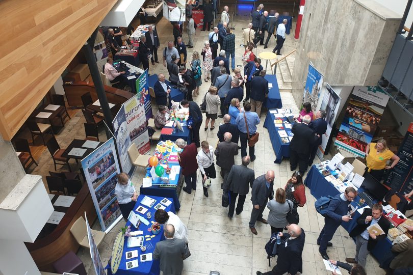 Overhead view of a busy indoor exhibition event at the Millennium Forum, with people engaging at various promotional stalls and information stands.
