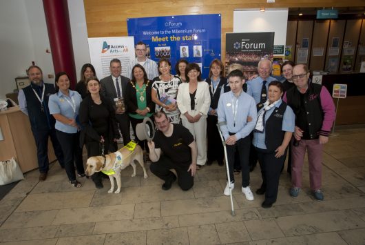 A group of people, including individuals with visual impairments, guide dogs, and Millennium Forum staff, pose together in the theatre's reception area in front of a “Meet the Staff” board and accessibility banners.