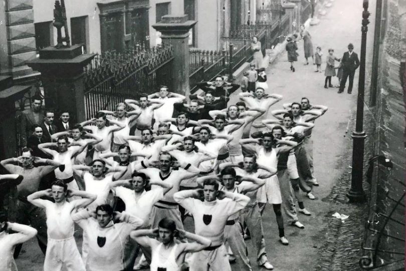 Black and white photo of a large group of young women and girls performing synchronised exercises or dance in a street in Derry, lined with onlookers and period buildings.
