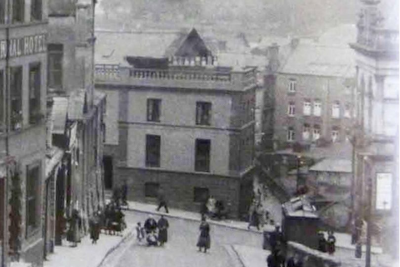 Historic black and white photo of a steep street in Derry, showing early 20th-century buildings, pedestrians in period clothing, and a tram or cart on the road.