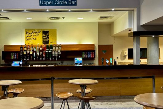 The Upper Piazza Bar at the Millennium Forum, featuring a wooden service counter with drinks dispensers, a till area, and a vibrant yellow wall sign displaying positive messages. In front of the bar are round wooden tables with matching stools for patrons.
