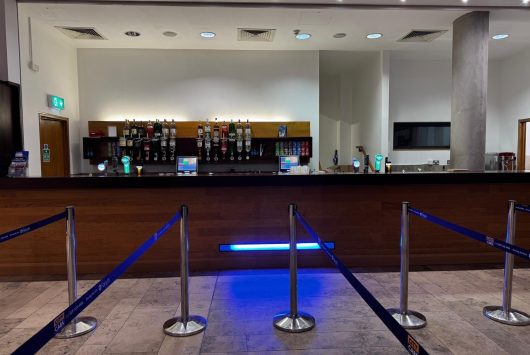 A sleek piazza bar with a wooden counter and a range of bottled and draught drinks on display. Blue LED lighting highlights the queue area, which is set up with stanchions and tape. The space features white walls, ceiling spotlights, and a polished tiled floor.