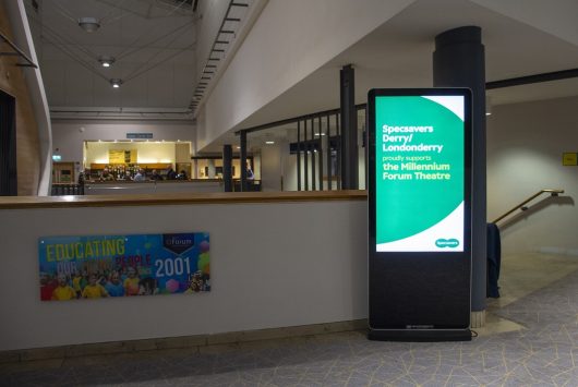 Digital signage inside the Millennium Forum displays a green Specsavers screen reading “Specsavers Derry/Londonderry supports the Millennium Forum Theatre,” beside a colourful mural and view of the foyer.