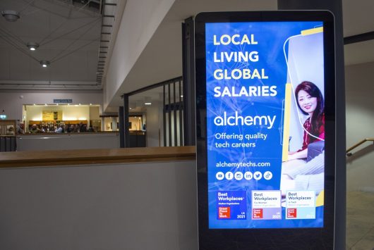 Digital screen inside the Millennium Forum displaying an Alchemy Technology Services advert with the text “Local living, global salaries” and a photo of a woman using a laptop.