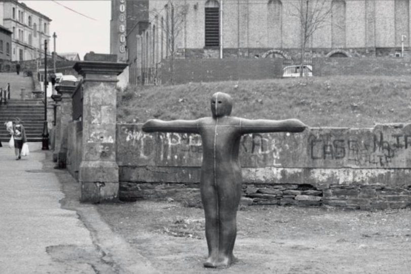 Black and white photo of a human-shaped sculpture with outstretched arms on a street in Derry, with worn stone walls and urban buildings in the background.