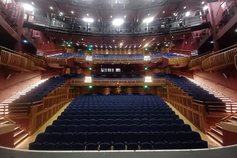 View of the Millennium Forum auditorium from the stage, showing rows of blue seats, tiered balconies, and warm lighting throughout the modern theatre interior.