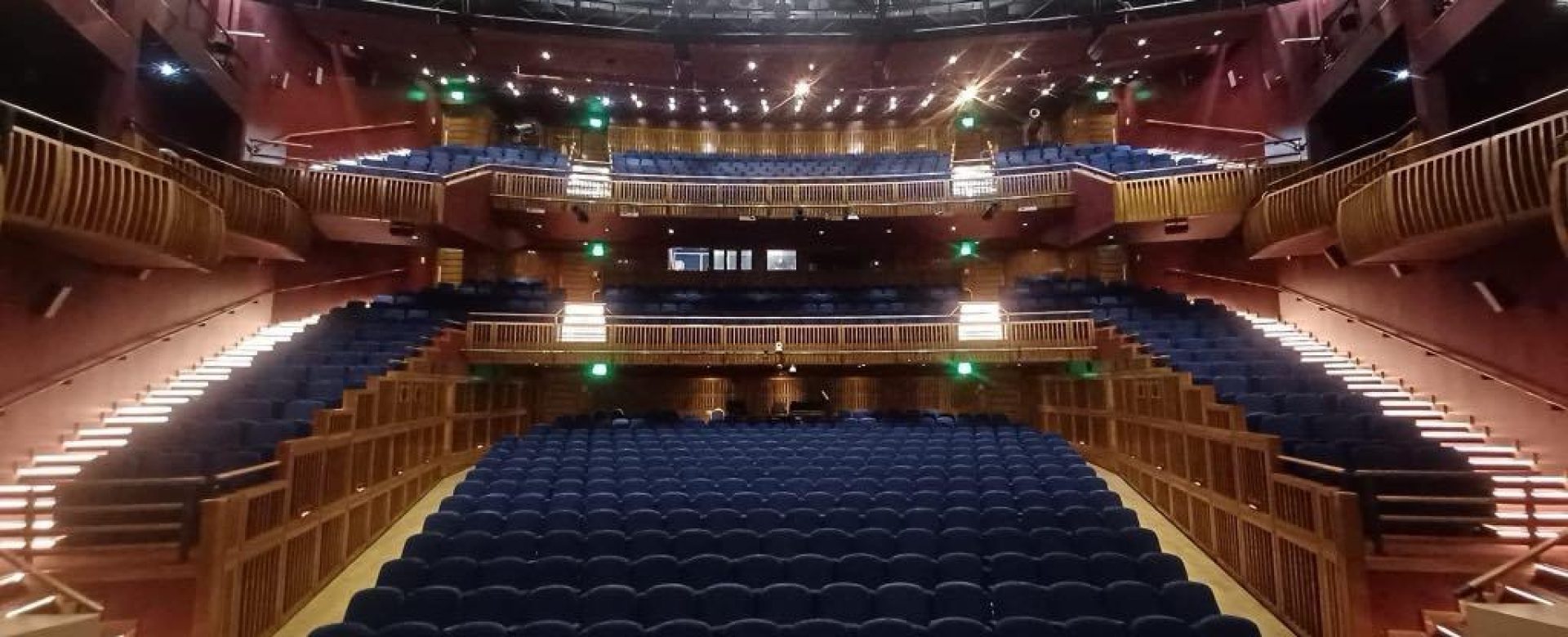 View of the Millennium Forum auditorium from the stage, showing rows of blue seats, tiered balconies, and warm lighting throughout the modern theatre interior.