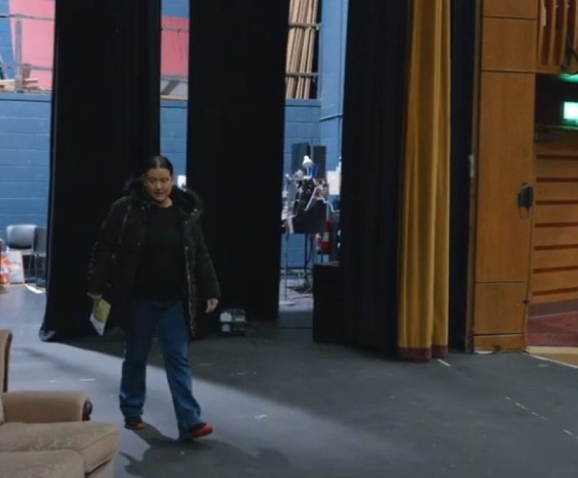 A person stands on stage at the Millennium Forum, near backstage curtains, with stage props and seating in the background. The wings and part of the auditorium are visible.