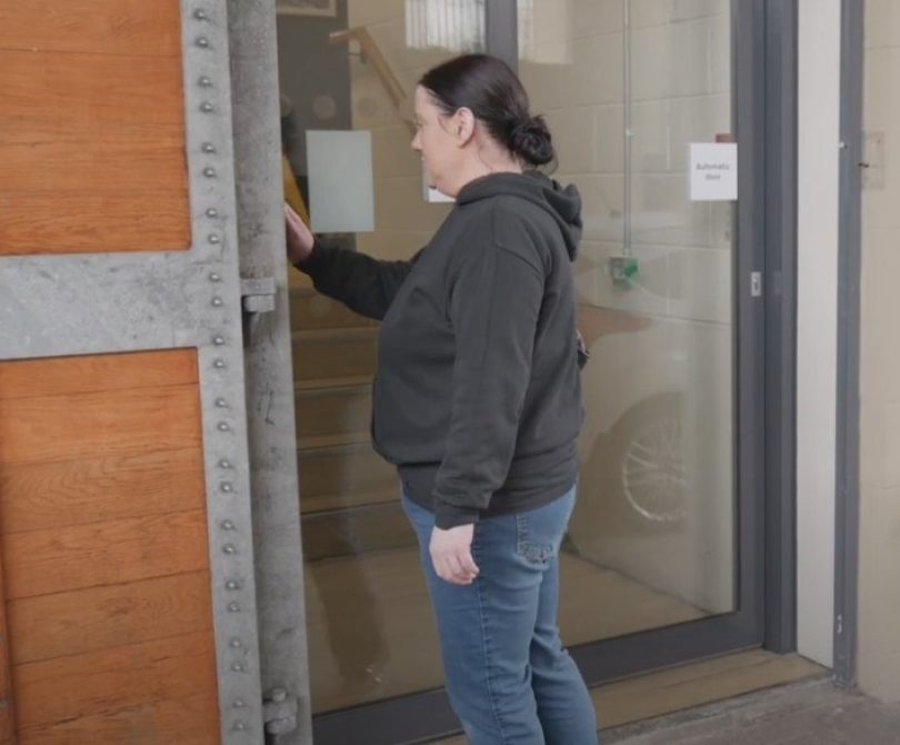 A person in casual clothing stands at an entrance door, reaching to press a buzzer beside a secure staff access point at the Millennium Forum.