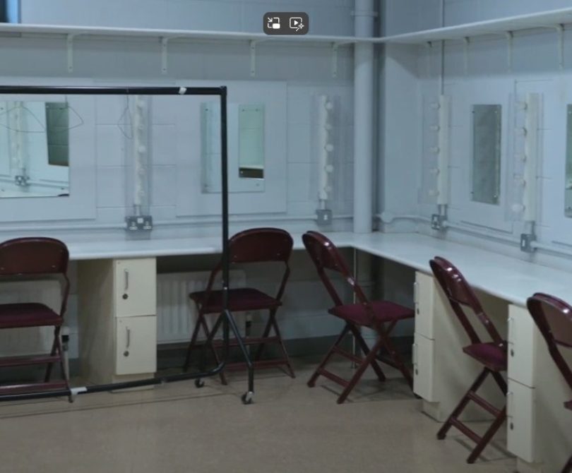 A dressing room with multiple vanity mirrors and lights, maroon folding chairs, white countertops, and lockers underneath. A costume rail stands in the centre of the room.