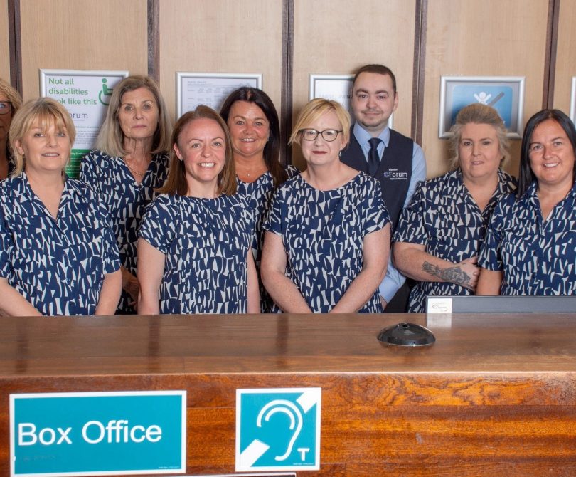 Millennium Forum Box Office team standing behind the counter, smiling, with Box Office and hearing assistance signs visible in front.