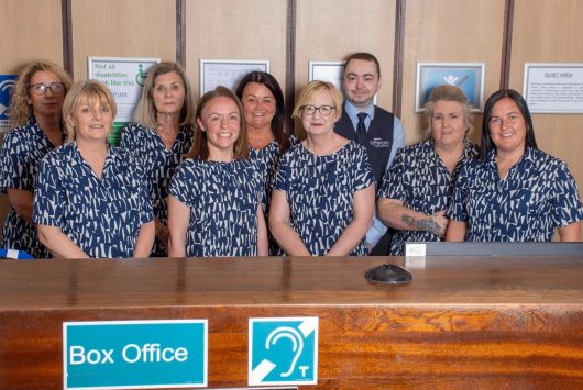 Millennium Forum Box Office team standing behind the counter, smiling, with Box Office and hearing assistance signs visible in front.