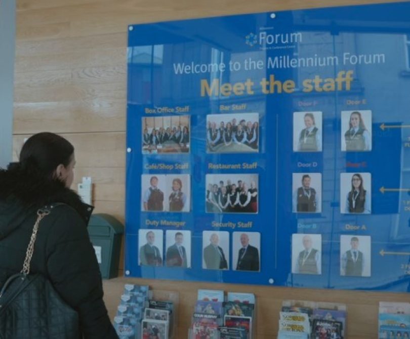 Video Thumbnail of a person standing in front of a large blue “Meet the Staff” board at the Millennium Forum, displaying staff photos and names. Leaflets and brochures are arranged on shelves below the board.