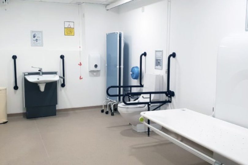 Accessible changing facility with hoist and adult-sized changing bench, equipped with a toilet, sink, grab rails, and privacy screen in a bright, hygienic environment.