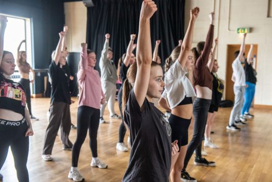A group of young people in a dance or drama workshop at the Millennium Forum, standing in rows with arms raised in unison inside a studio space with wooden floors.