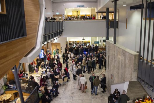 A busy theatre foyer filled with people socialising and queuing at the café bar area, with a modern interior featuring marble floors, balcony railings, and signage for upcoming performances.