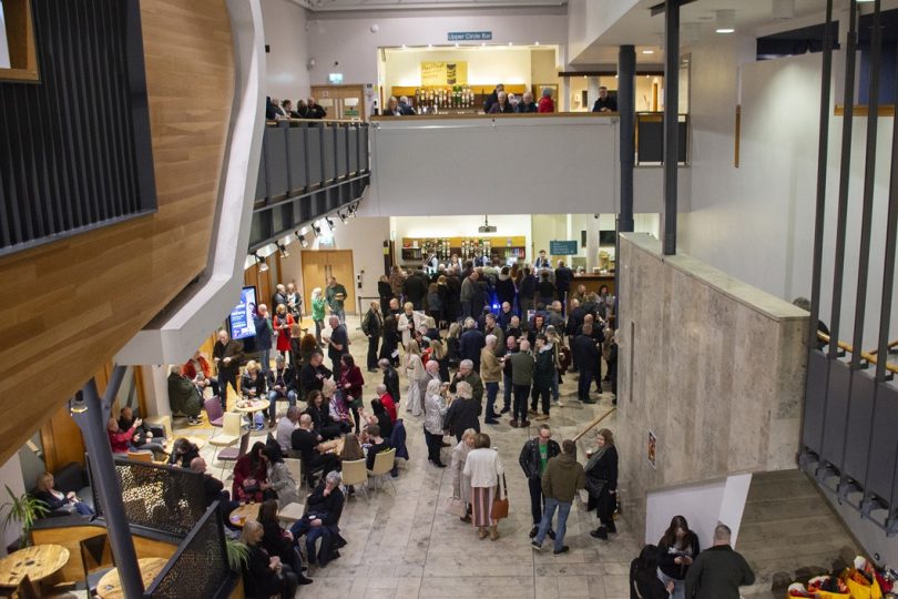 A busy theatre foyer filled with people socialising and queuing at the café bar area, with a modern interior featuring marble floors, balcony railings, and signage for upcoming performances.