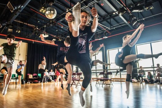 Group of young dancers mid-air during a high-energy jump in a rehearsal studio, with sunlight streaming through the windows and a mirrored disco ball above.