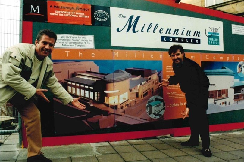 Two men smiling and pointing to a construction hoarding featuring a large design visual for the Millennium Forum theatre, with branding and project details displayed.