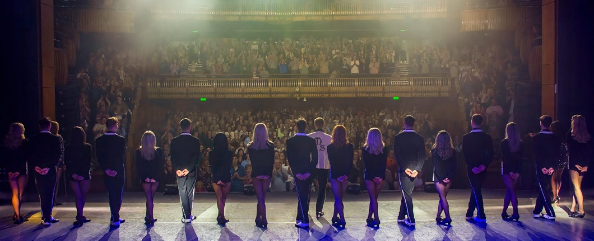A cast of performers from Lord of the Dance stands in a line facing the audience for a final bow on stage at the Millennium Forum, with bright stage lights and a packed auditorium in view.