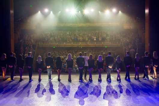 A cast of performers from Lord of the Dance stands in a line facing the audience for a final bow on stage at the Millennium Forum, with bright stage lights and a packed auditorium in view.
