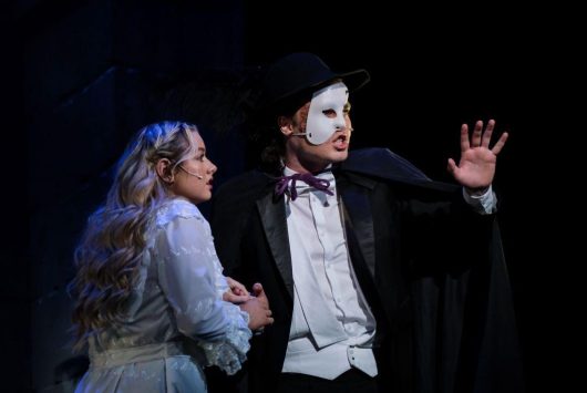 Two actors on stage in costume during a dramatic scene: a man wearing a white half-mask, cape, and top hat holds up his hand while a woman in a white dress stands beside him, looking concerned.
