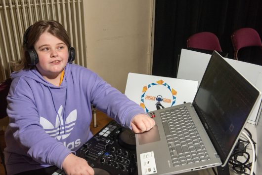 A young person in a purple hoodie wearing headphones operates DJ equipment and a laptop during a music or tech workshop.