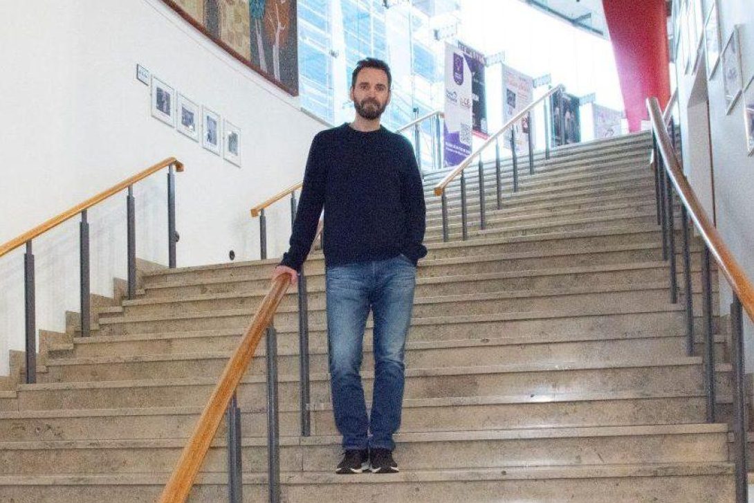 Johnny McDaid stands on the wide staircase inside the Millennium Forum, surrounded by poster displays and gallery lighting.