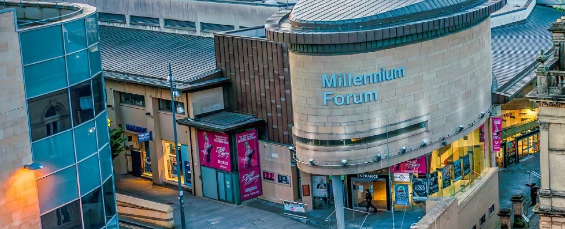 Exterior view of the Millennium Forum theatre in Derry, showing the curved modern architecture, main entrance, and colourful show posters on display. The image is taken from an elevated angle, capturing surrounding buildings and the cityscape in the background.