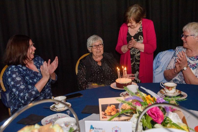 A group of older women gathered around a table, smiling and clapping as one of them is presented with a small birthday cake topped with lit candles during a tea party celebration.