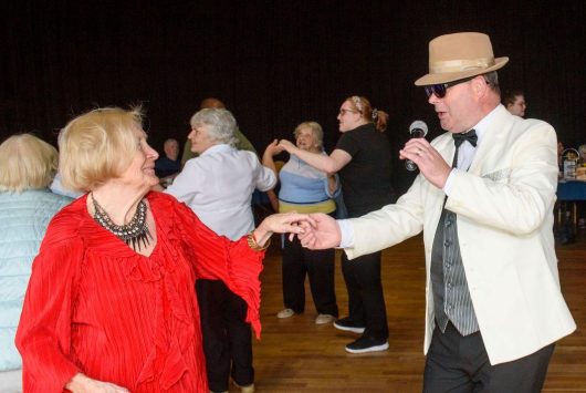 A singer in a cream jacket and hat holds a microphone while dancing with an older woman in a red top at a community tea dance, with other guests dancing in the background.