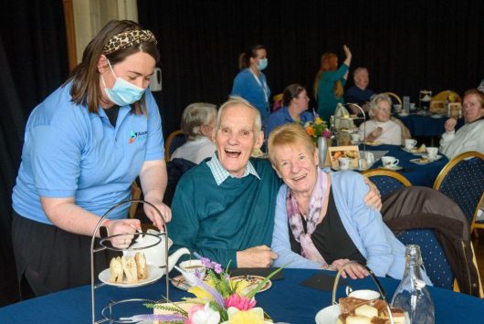 An older couple smiling and holding hands at a dementia-friendly afternoon tea event, as a staff member serves sandwiches. Other guests and carers are seated in the background.