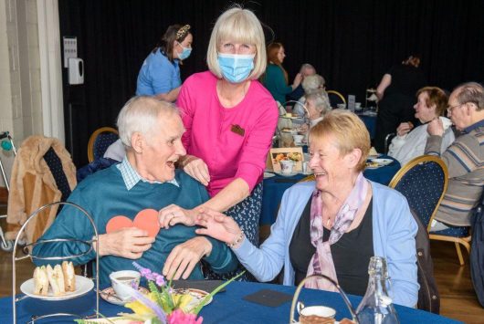 A smiling woman wearing a mask engages warmly with an older couple seated at a table during a tea dance or community event, with afternoon tea stands and flowers on the table.