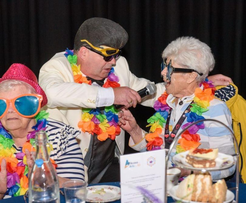 Two older women in colourful hats and oversized sunglasses enjoy a fun moment as a performer in costume sings into a microphone at their table during a dementia-friendly tea dance.