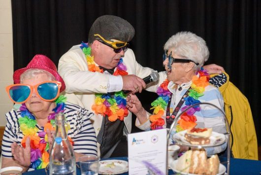 Two older women in colourful hats and oversized sunglasses enjoy a fun moment as a performer in costume sings into a microphone at their table during a dementia-friendly tea dance.