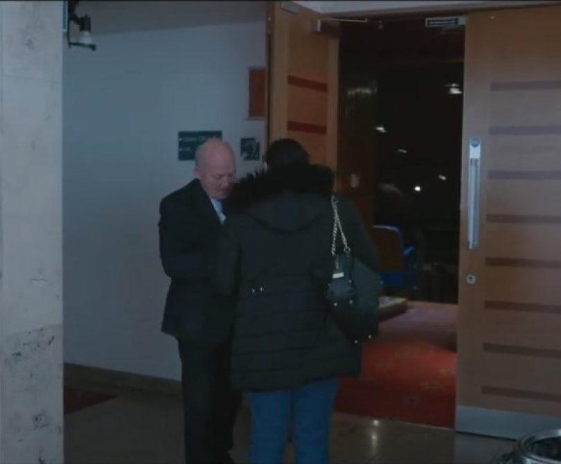 A woman in a black coat is greeted by an usher at the entrance to a theatre auditorium inside the Millennium Forum, with wooden double doors open behind them.