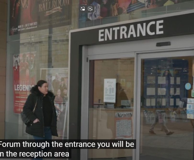 A woman approaches the main entrance of the Millennium Forum, where large glass doors are labelled “ENTRANCE.” Posters for upcoming shows are visible through the glass walls.