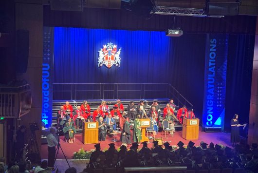 Ulster University graduation ceremony on stage at the Millennium Forum, with graduates in caps and gowns seated before a panel of faculty members in red academic robes, and large blue banners reading "#PROUDofUU" and "CONGRATULATIONS".