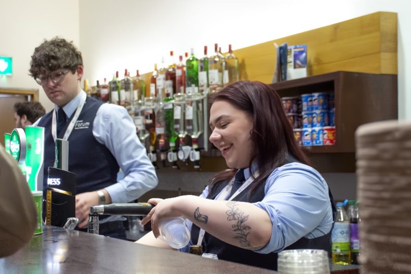 Two smiling Millennium Forum bar staff behind the counter, serving drinks with shelves of spirits and mixers in the background.