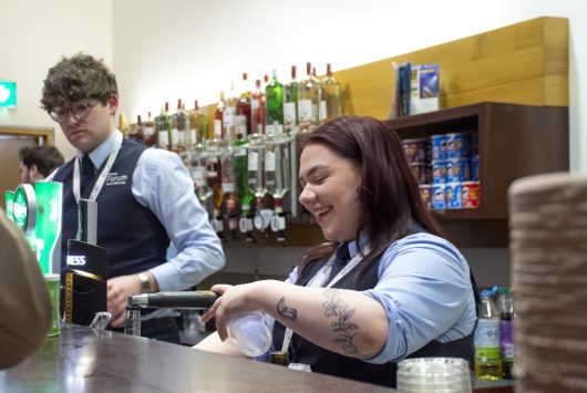 Two smiling Millennium Forum bar staff behind the counter, serving drinks with shelves of spirits and mixers in the background.