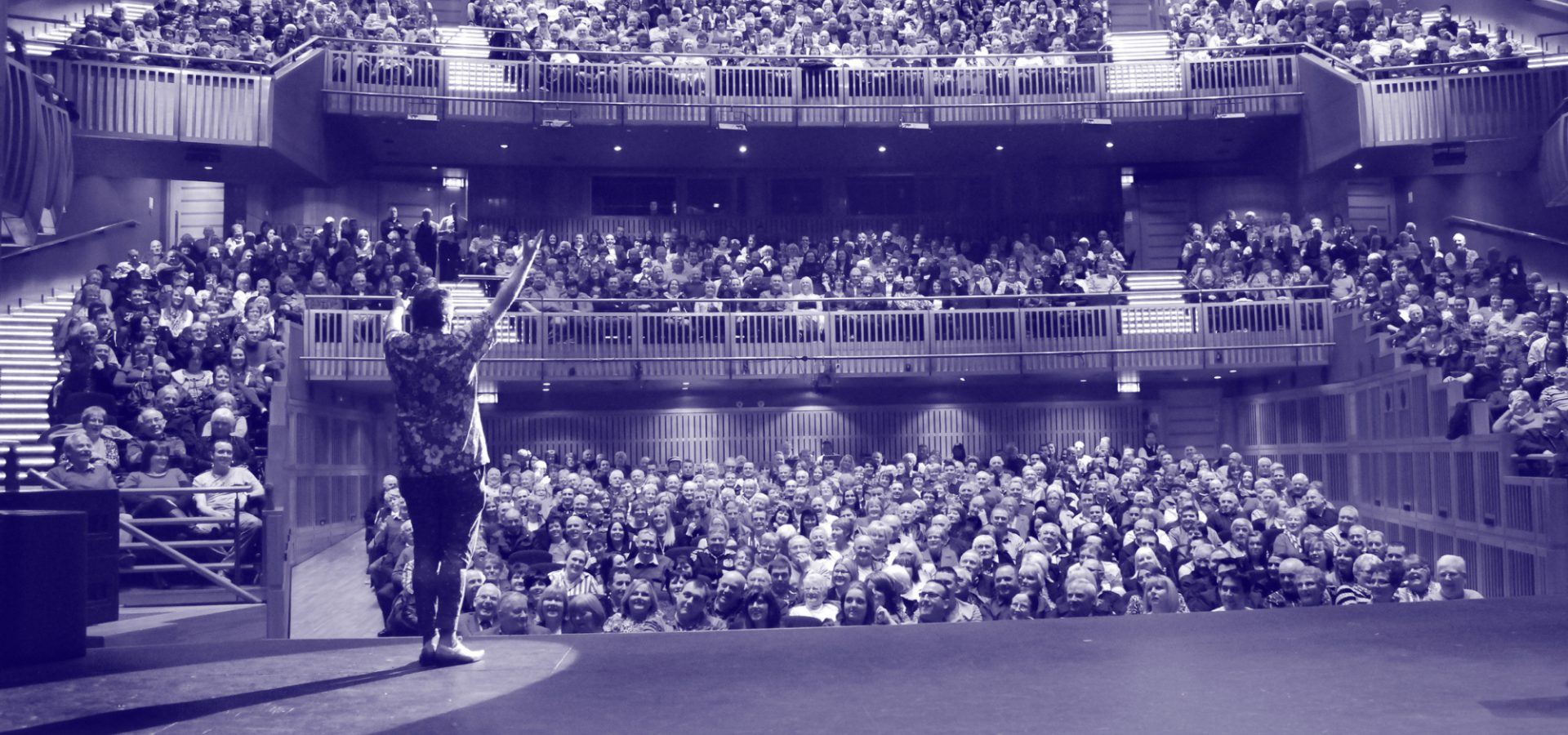 View from the stage of a packed auditorium at the Millennium Forum, showing a performer addressing a full house across all seating levels. The image has a purple-blue overlay, creating a bold and dynamic backdrop