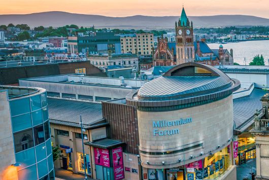 Wide-angle view of the Millennium Forum in Derry, Northern Ireland, adjacent to the iconic Derry Girls mural. The scene includes historic buildings and a clear blue sky, blending modern culture with local heritage.