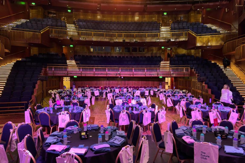 Millennium Forum auditorium transformed for a corporate event with round tables, chairs draped in white gift bags, and purple lighting highlighting the stage and seating tiers.