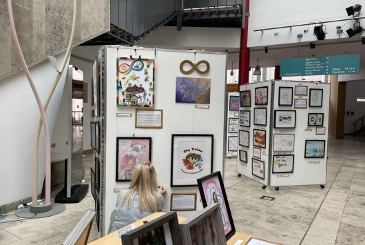 Art exhibition in the Millennium Forum foyer displaying framed artwork on white panels. A visitor is seated viewing the artwork, with natural light and modern architectural features visible in the space.