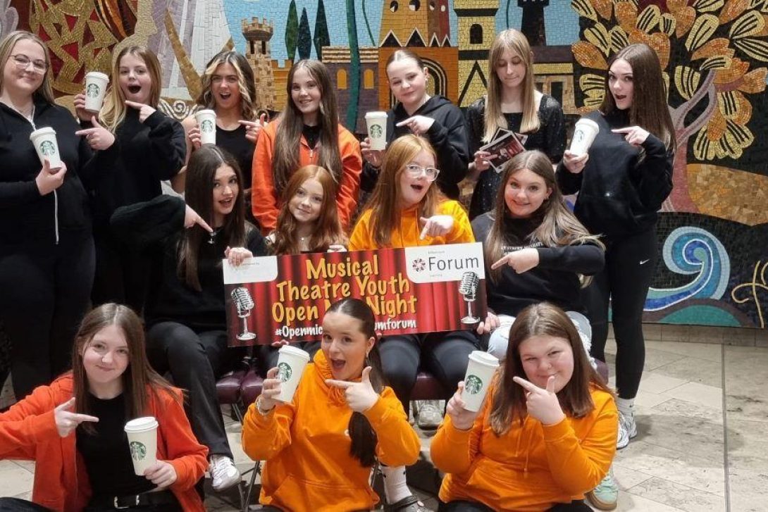 "Group of smiling teenage girls posing in front of a vibrant mural at the Millennium Forum, holding Starbucks cups and pointing at a red and gold sign that reads 'Musical Theatre Youth Open Night @Forum'. Several wear orange Forum hoodies, suggesting involvement in a youth arts programme.
