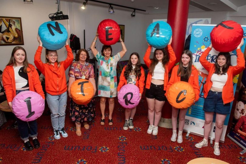 A group of young people in orange hoodies and two adults stand in a line, each holding a large colourful balloon with a letter spelling out “INTERACT”, during a youth event at the Millennium Forum.
